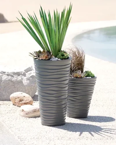 Donica Sand Slim 75cm mocca w aranżacji z zielonymi roślinami ozdobnymi, stojąca na piasku.