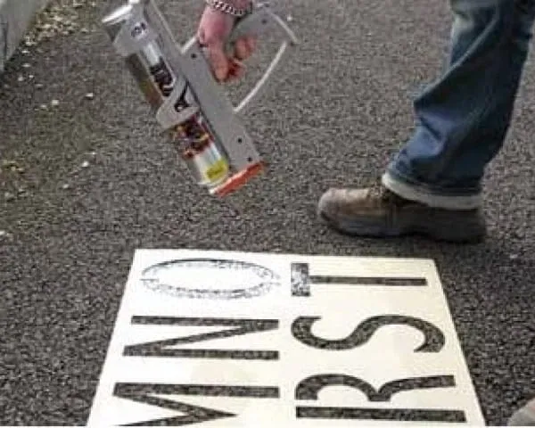 Pracownik malujący napis na ziemi za pomocą pistoletu ręcznego i metalowego szablonu.