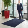 Niebieska wycieraczka obiektowa Monokolor 85x60 cm położona w strefie wejściowej biurowca z automatycznymi drzwiami i mężczyzną przechodzącym po nylonowym runie.